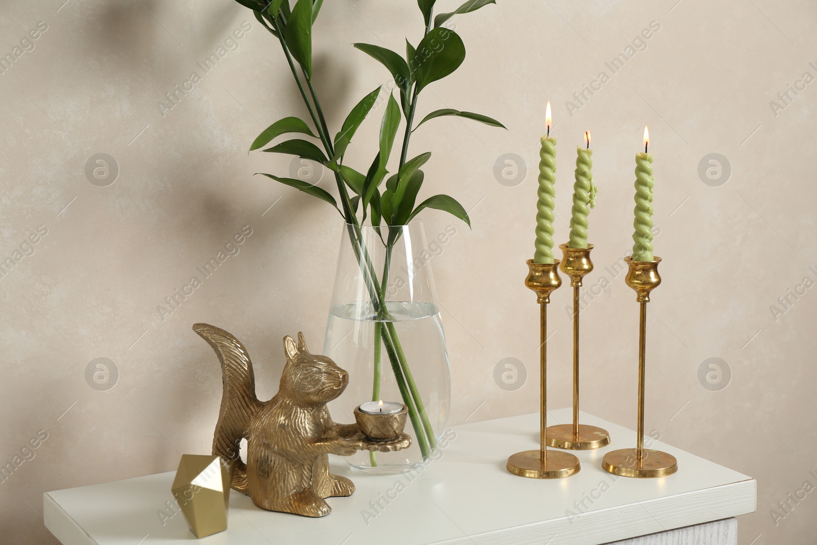 Elegant candlesticks with burning candles, branches in vase and other decor on dresser near beige wall Photo of Elegant candlesticks with burning candles, branches in vase and other decor on dresser near beige wall