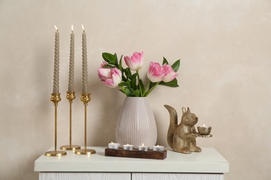 Photo of Elegant candlesticks with burning candles, tulips in vase and other decor on dresser near beige wall