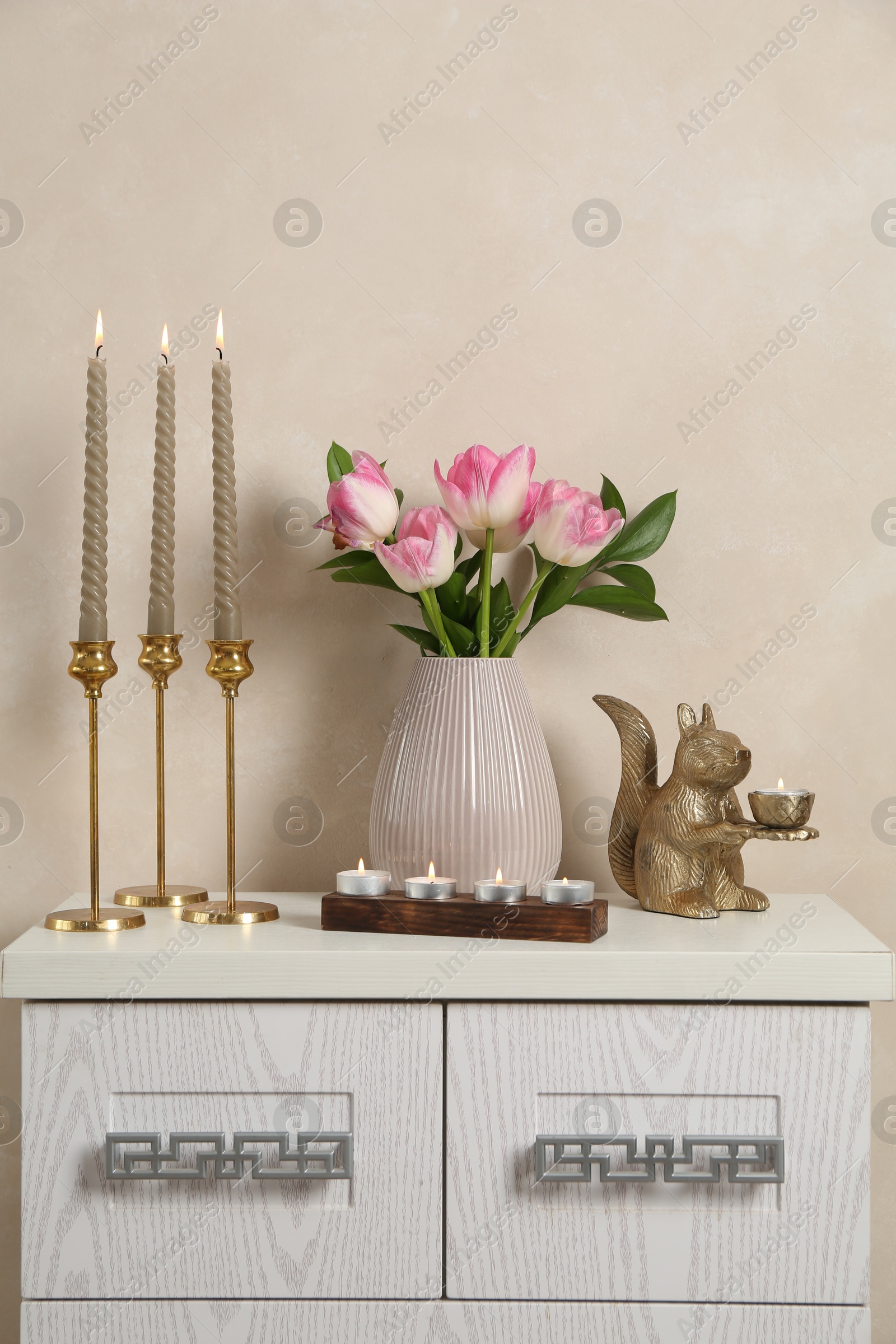 Elegant candlesticks with burning candles, tulips in vase and other decor on dresser near beige wall Photo of Elegant candlesticks with burning candles, tulips in vase and other decor on dresser near beige wall