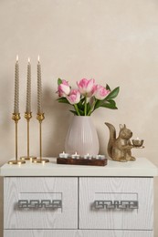Photo of Elegant candlesticks with burning candles, tulips in vase and other decor on dresser near beige wall