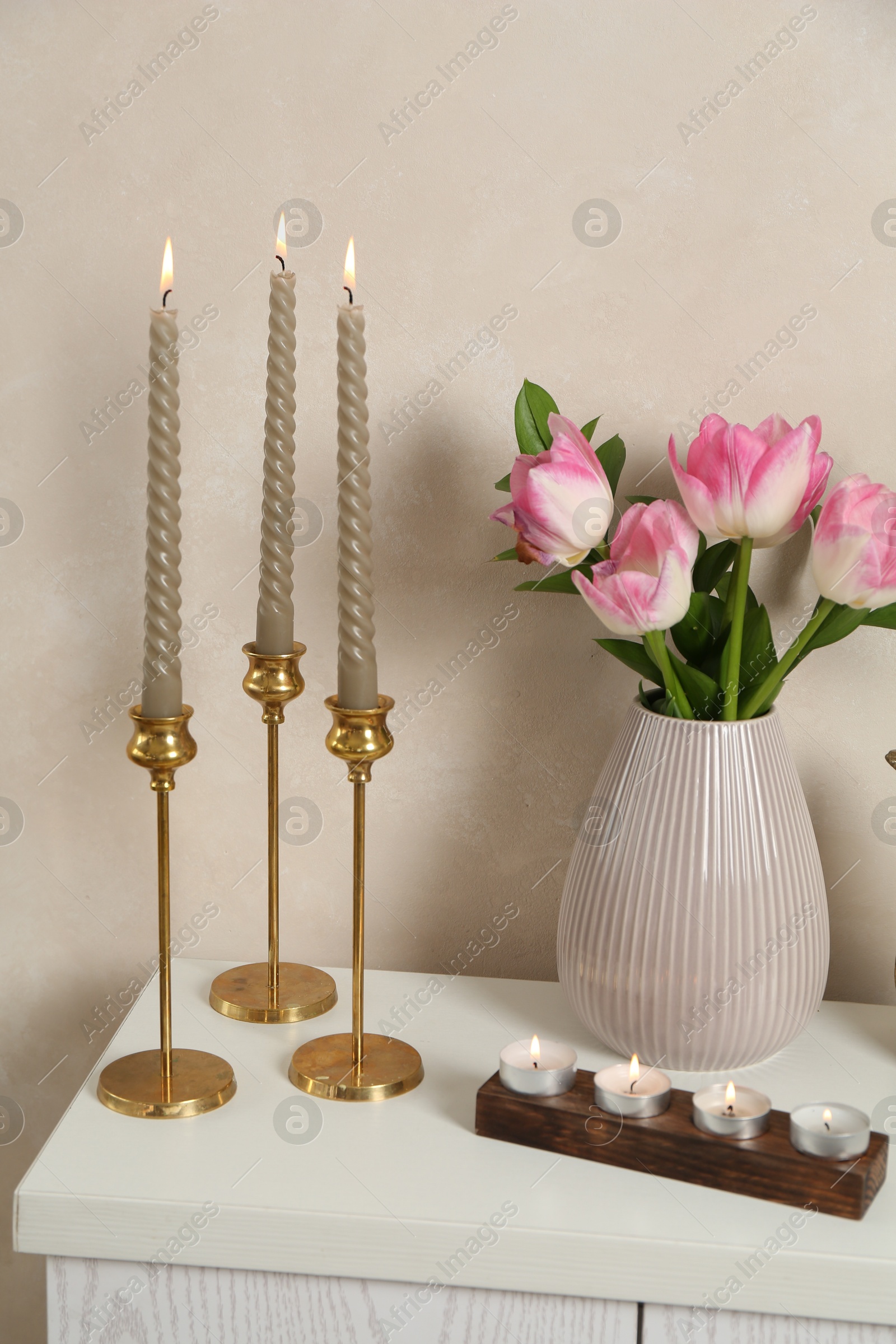 Elegant candlesticks with burning candles and tulips in vase on dresser near beige wall Photo of Elegant candlesticks with burning candles and tulips in vase on dresser near beige wall