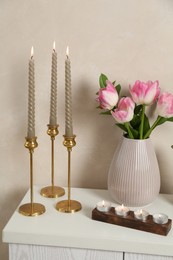 Photo of Elegant candlesticks with burning candles and tulips in vase on dresser near beige wall