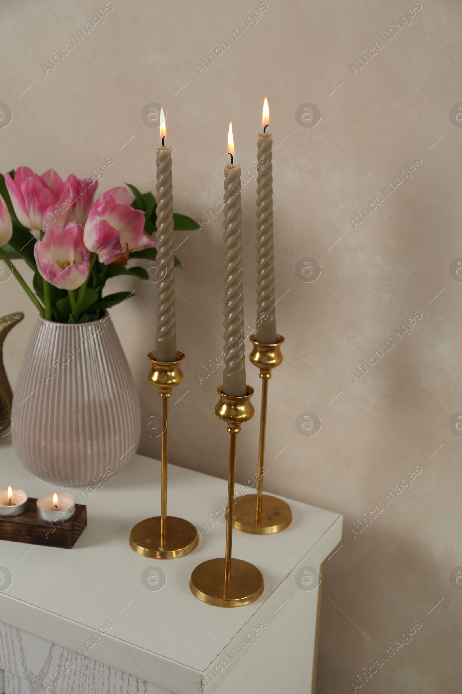 Elegant candlesticks with burning candles and tulips in vase on dresser near beige wall Photo of Elegant candlesticks with burning candles and tulips in vase on dresser near beige wall