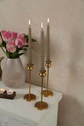 Photo of Elegant candlesticks with burning candles and tulips in vase on dresser near beige wall