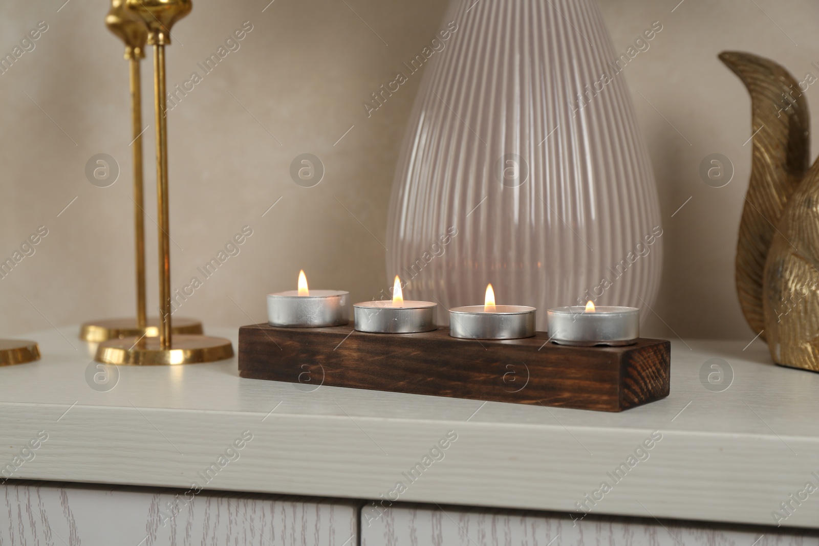 Elegant holders with burning candles and decor on dresser near beige wall, closeup Photo of Elegant holders with burning candles and decor on dresser near beige wall, closeup