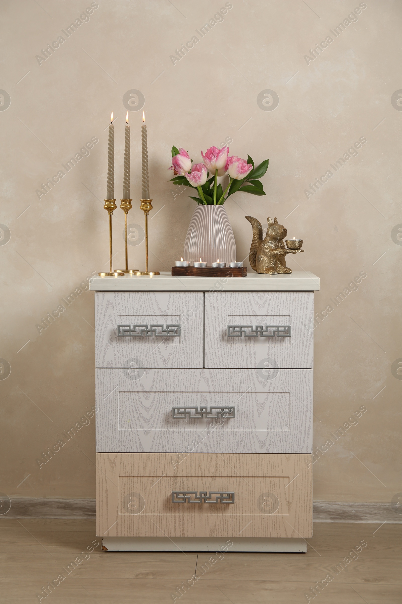 Elegant candlesticks with burning candles, tulips in vase and other decor on dresser near beige wall Photo of Elegant candlesticks with burning candles, tulips in vase and other decor on dresser near beige wall