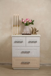 Photo of Elegant candlesticks with burning candles, tulips in vase and other decor on dresser near beige wall
