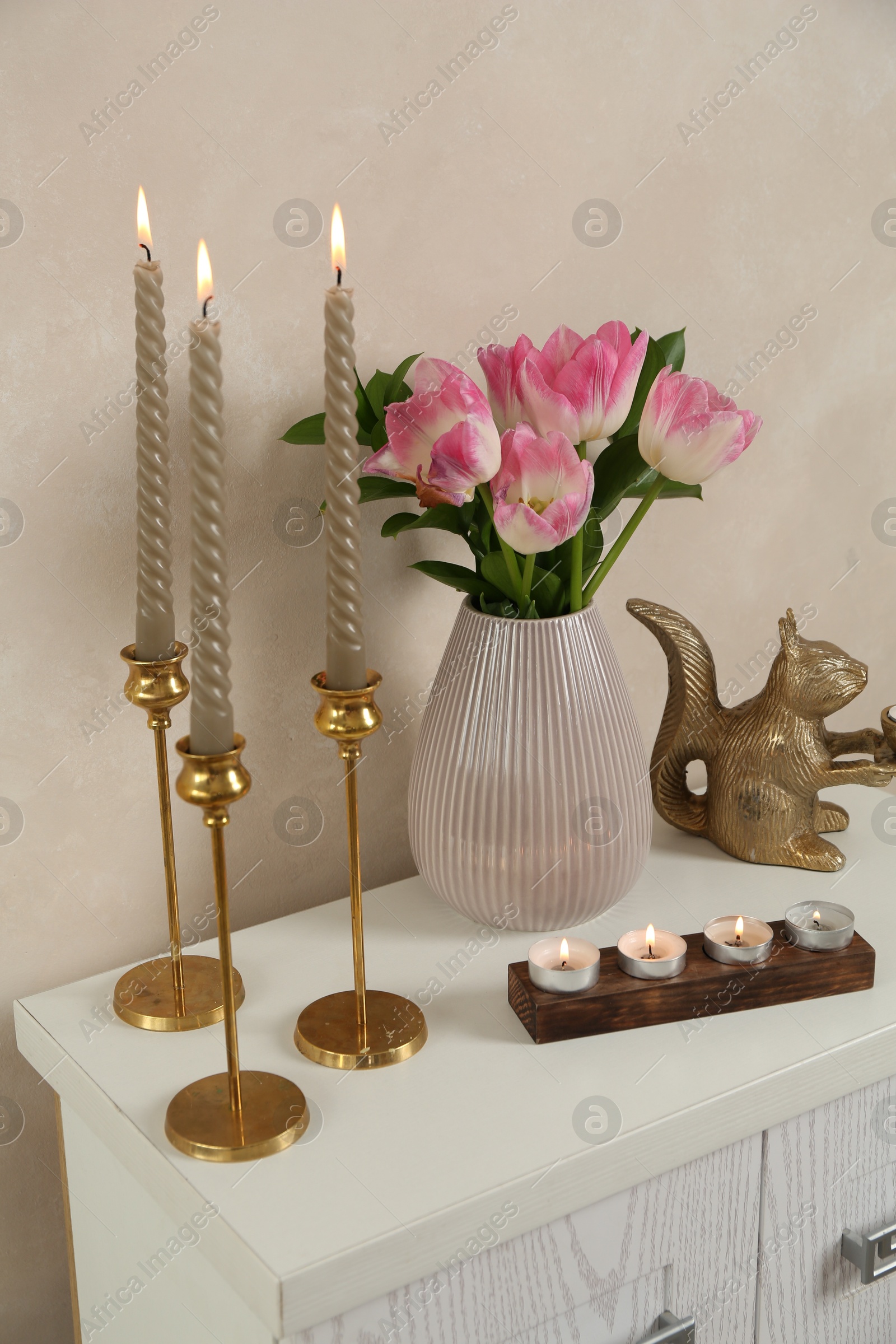 Elegant candlesticks with burning candles, tulips in vase and other decor on dresser near beige wall Photo of Elegant candlesticks with burning candles, tulips in vase and other decor on dresser near beige wall