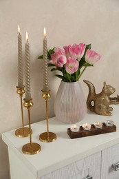 Photo of Elegant candlesticks with burning candles, tulips in vase and other decor on dresser near beige wall