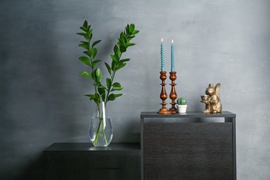 Photo of Elegant candlesticks with burning candles, branches in vase and other decor on dresser near grey wall