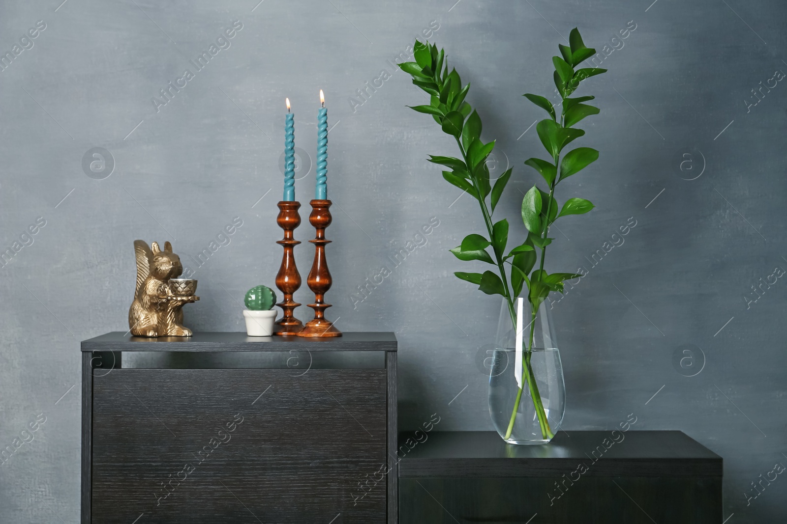 Elegant candlesticks with burning candles, branches in vase and other decor on dresser near grey wall Photo of Elegant candlesticks with burning candles, branches in vase and other decor on dresser near grey wall