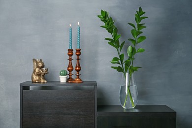 Photo of Elegant candlesticks with burning candles, branches in vase and other decor on dresser near grey wall