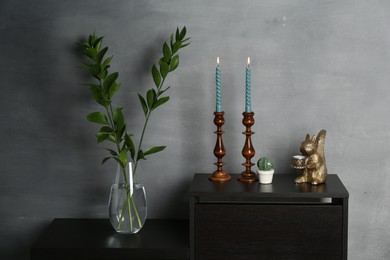 Photo of Elegant candlesticks with burning candles, branches in vase and other decor on dresser near grey wall