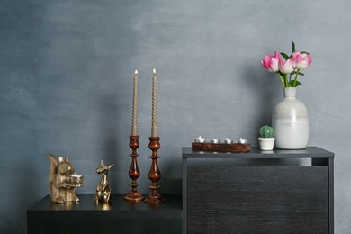 Photo of Elegant candlesticks with burning candles, tulips in vase and other decor on dresser near grey wall