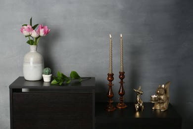 Photo of Elegant candlesticks with burning candles, tulips in vase and other decor on dresser near grey wall