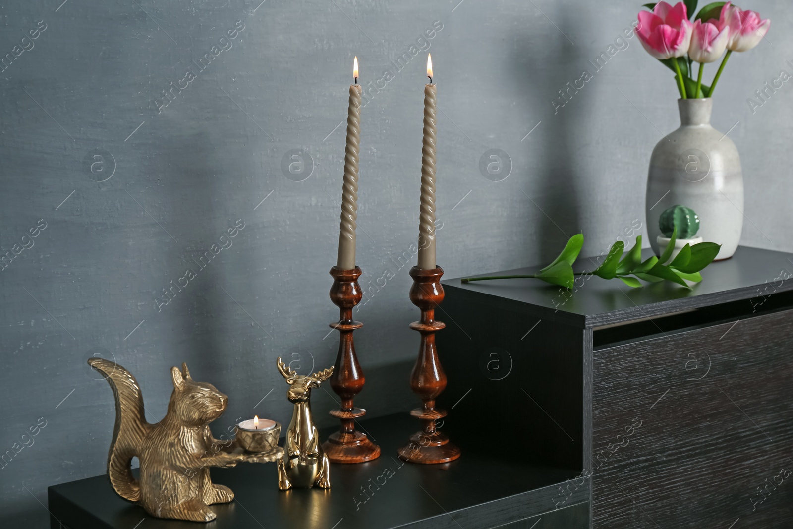 Elegant candlesticks with burning candles, tulips in vase and other decor on dresser near grey wall Photo of Elegant candlesticks with burning candles, tulips in vase and other decor on dresser near grey wall