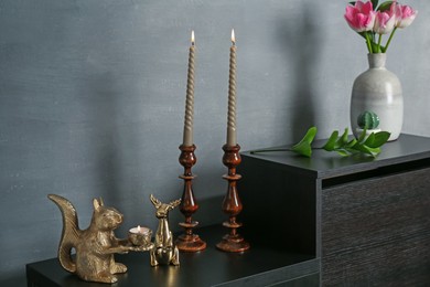 Photo of Elegant candlesticks with burning candles, tulips in vase and other decor on dresser near grey wall