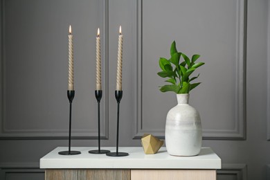 Photo of Elegant candlesticks with burning candles, branches in vase and other decor on dresser near grey wall