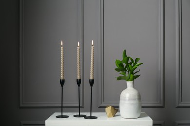 Photo of Elegant candlesticks with burning candles, branches in vase and other decor on dresser near grey wall