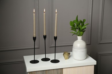 Photo of Elegant candlesticks with burning candles, branches in vase and other decor on dresser near grey wall