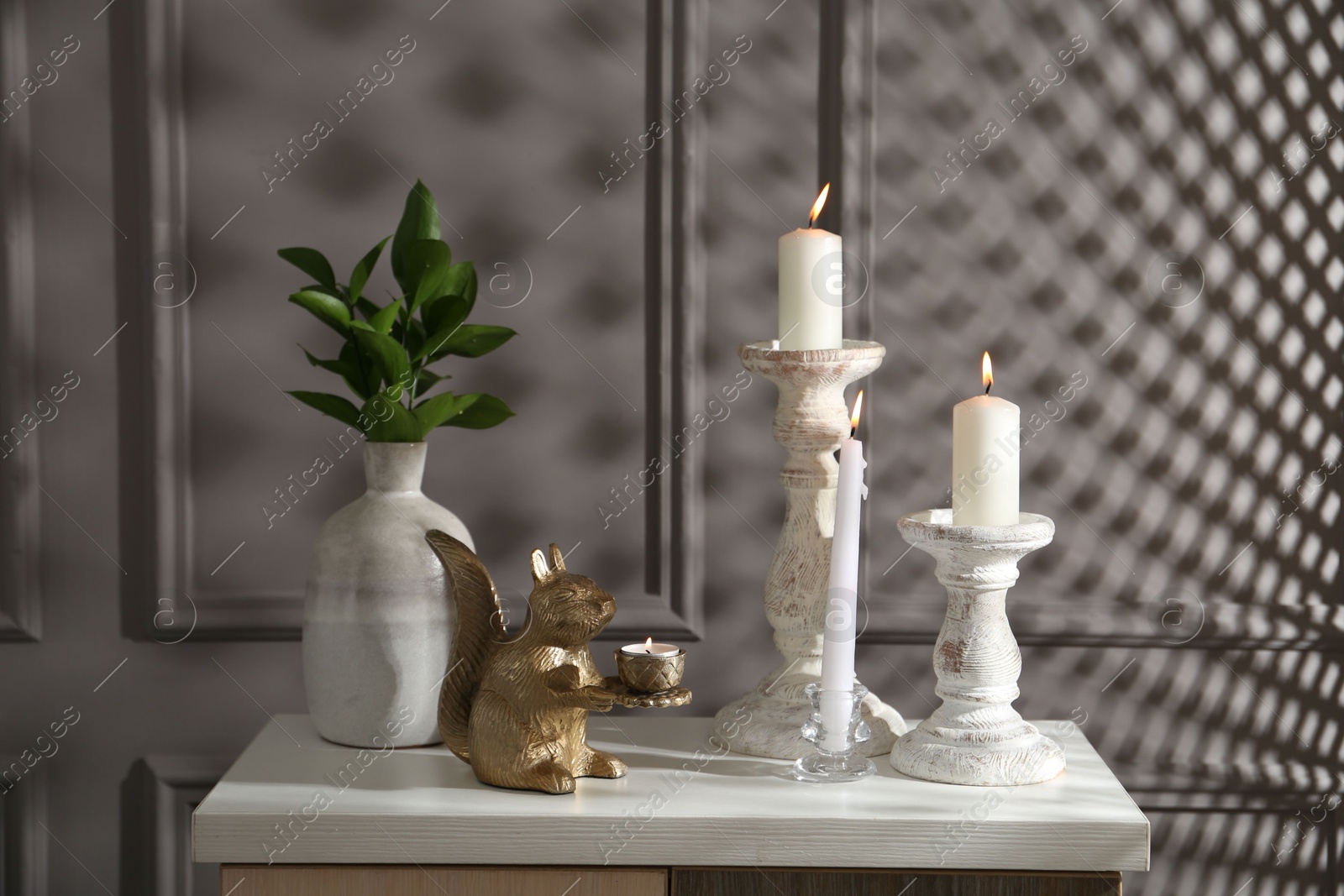 Elegant candlesticks with burning candles, branches in vase and other decor on dresser near grey wall Photo of Elegant candlesticks with burning candles, branches in vase and other decor on dresser near grey wall