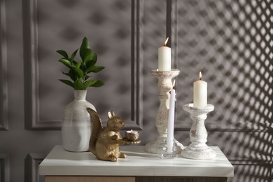 Photo of Elegant candlesticks with burning candles, branches in vase and other decor on dresser near grey wall