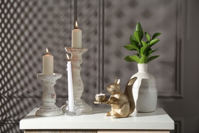 Photo of Elegant candlesticks with burning candles, branches in vase and other decor on dresser near grey wall