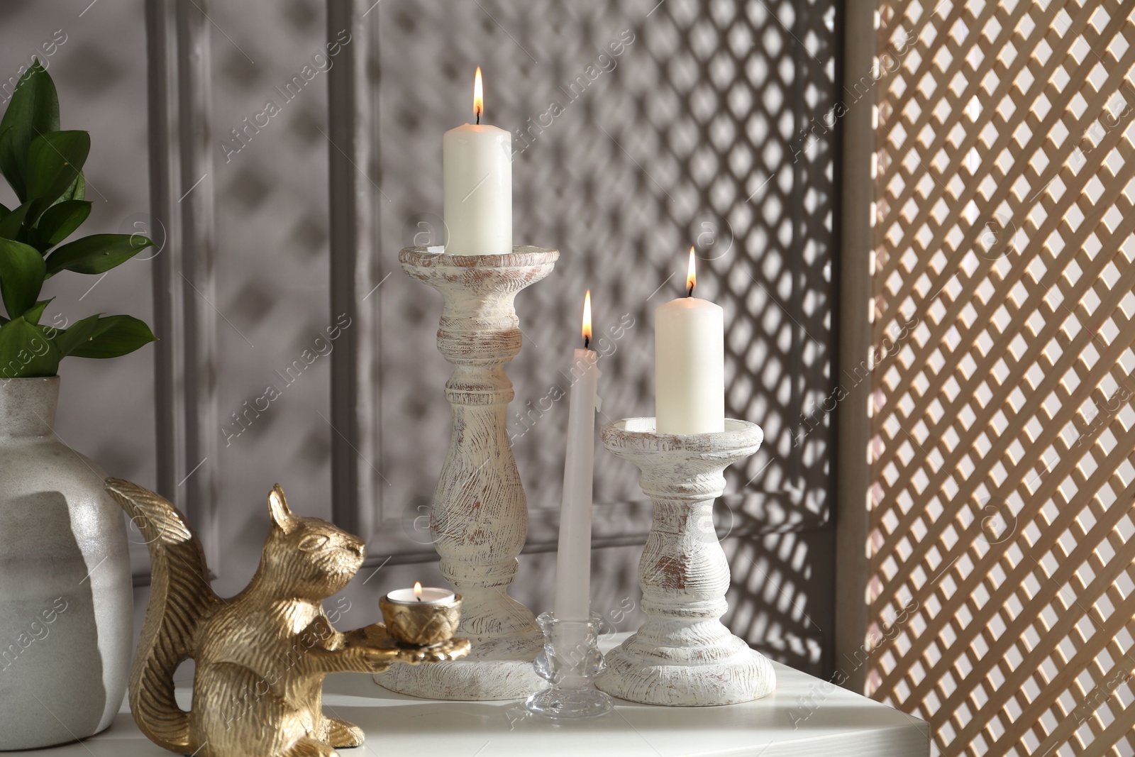 Elegant candlesticks with burning candles, branches in vase and other decor on dresser near grey wall Photo of Elegant candlesticks with burning candles, branches in vase and other decor on dresser near grey wall