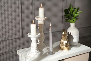 Photo of Elegant candlesticks with burning candles, branches in vase and other decor on dresser near grey wall