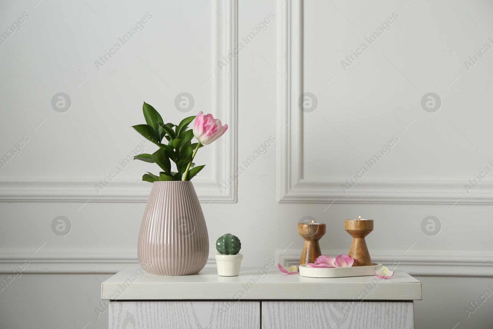 Elegant candlesticks with burning candles, tulip in vase and other decor on dresser near white wall Photo of Elegant candlesticks with burning candles, tulip in vase and other decor on dresser near white wall