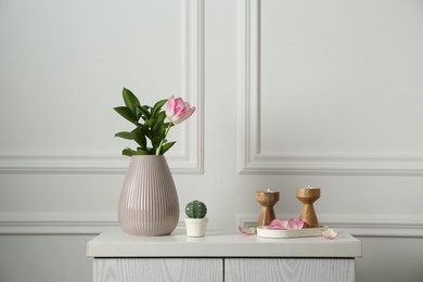Photo of Elegant candlesticks with burning candles, tulip in vase and other decor on dresser near white wall