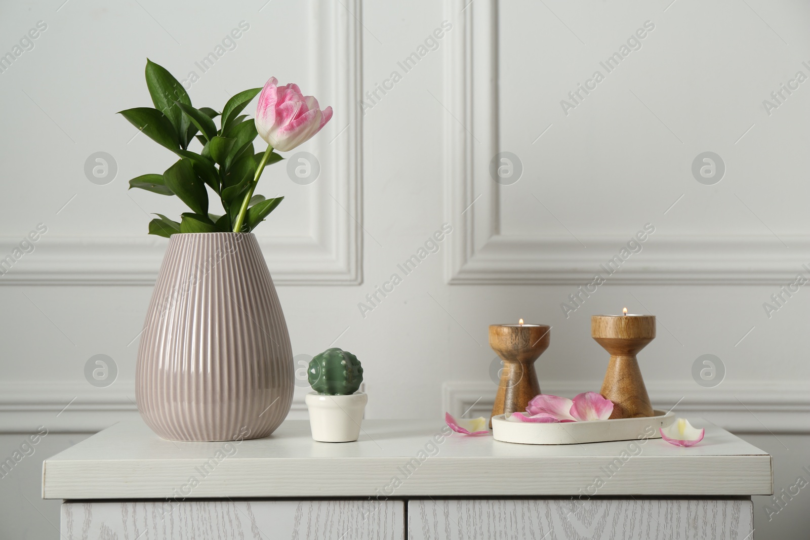 Elegant candlesticks with burning candles, tulip in vase and other decor on dresser near white wall Photo of Elegant candlesticks with burning candles, tulip in vase and other decor on dresser near white wall