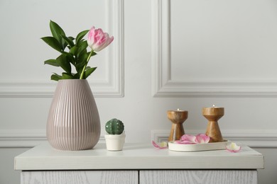 Photo of Elegant candlesticks with burning candles, tulip in vase and other decor on dresser near white wall