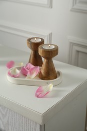 Photo of Elegant candlesticks with burning candles and flower petals on dresser near white wall, closeup
