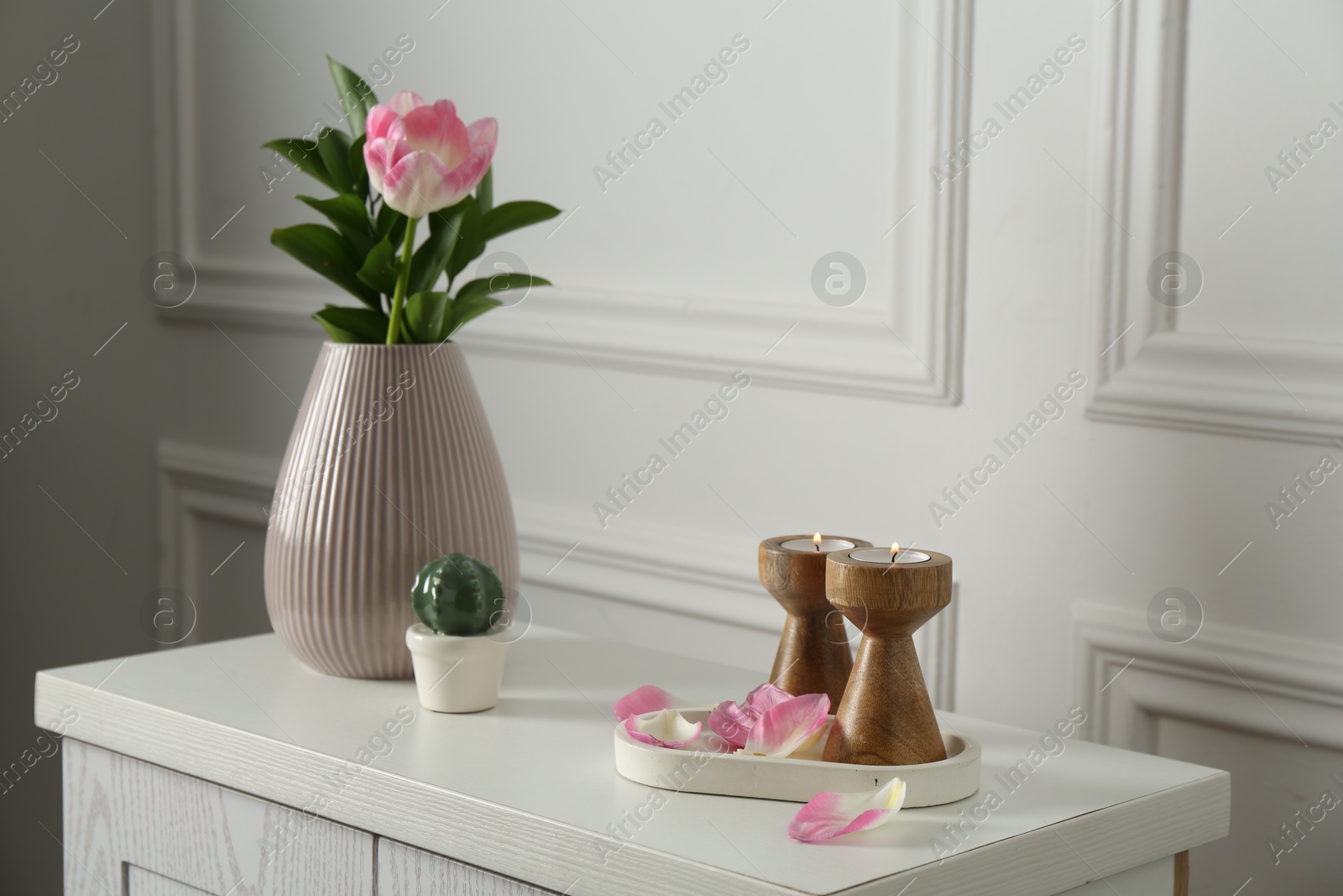 Elegant candlesticks with burning candles, tulip in vase and other decor on dresser near white wall Photo of Elegant candlesticks with burning candles, tulip in vase and other decor on dresser near white wall