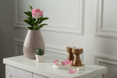 Photo of Elegant candlesticks with burning candles, tulip in vase and other decor on dresser near white wall