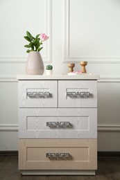 Photo of Elegant candlesticks with burning candles, tulip in vase and other decor on dresser near white wall