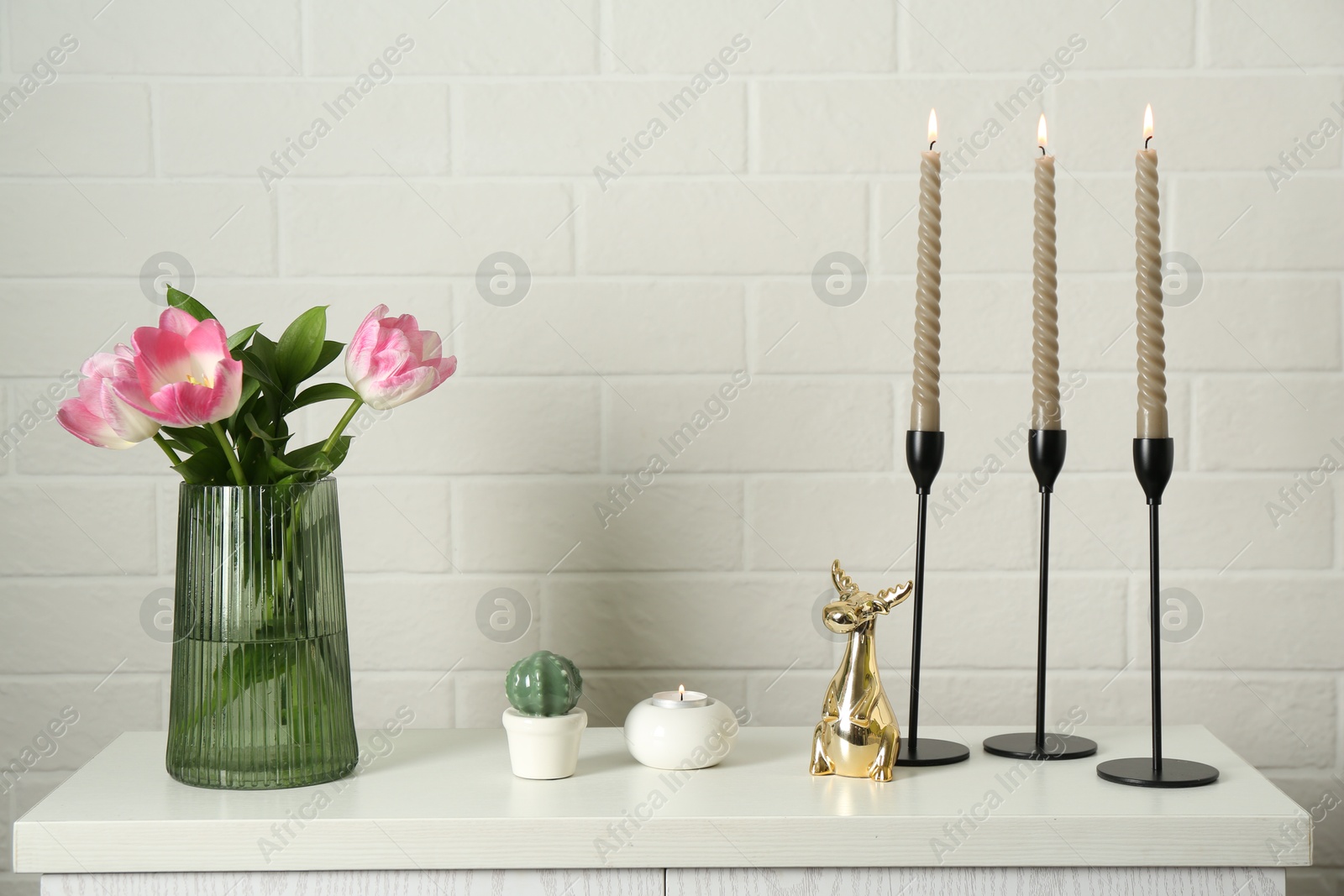 Elegant candlesticks with burning candles, tulips in vase and other decor on dresser near white brick wall Photo of Elegant candlesticks with burning candles, tulips in vase and other decor on dresser near white brick wall