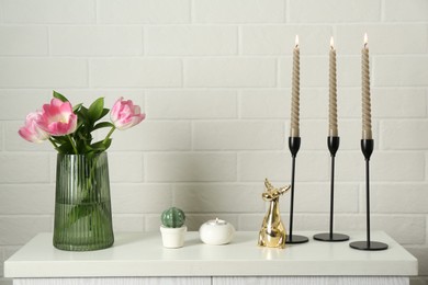 Photo of Elegant candlesticks with burning candles, tulips in vase and other decor on dresser near white brick wall