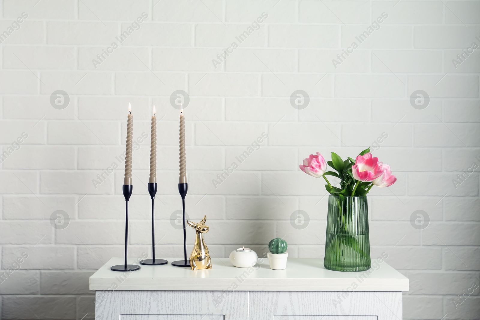 Elegant candlesticks with burning candles, tulips in vase and other decor on dresser near white brick wall Photo of Elegant candlesticks with burning candles, tulips in vase and other decor on dresser near white brick wall