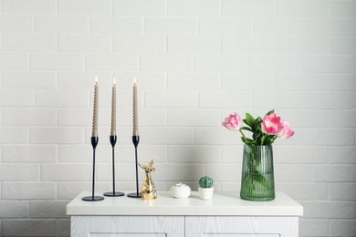 Photo of Elegant candlesticks with burning candles, tulips in vase and other decor on dresser near white brick wall