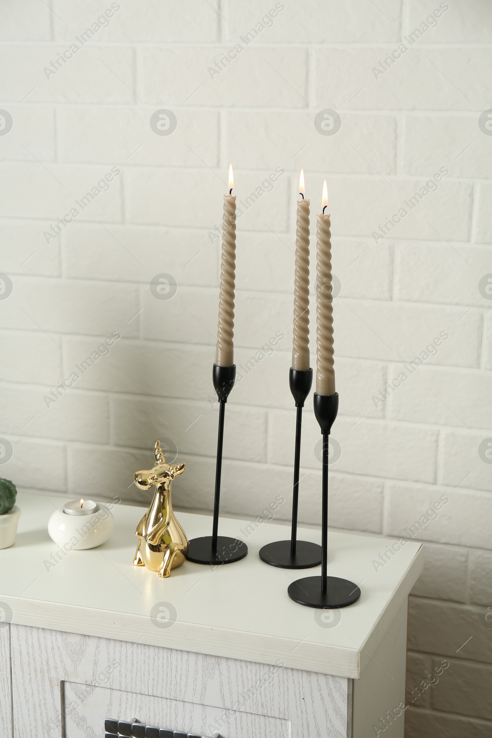 Elegant candlesticks with burning candles and other decor on dresser near white brick wall Photo of Elegant candlesticks with burning candles and other decor on dresser near white brick wall