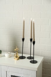 Photo of Elegant candlesticks with burning candles and other decor on dresser near white brick wall