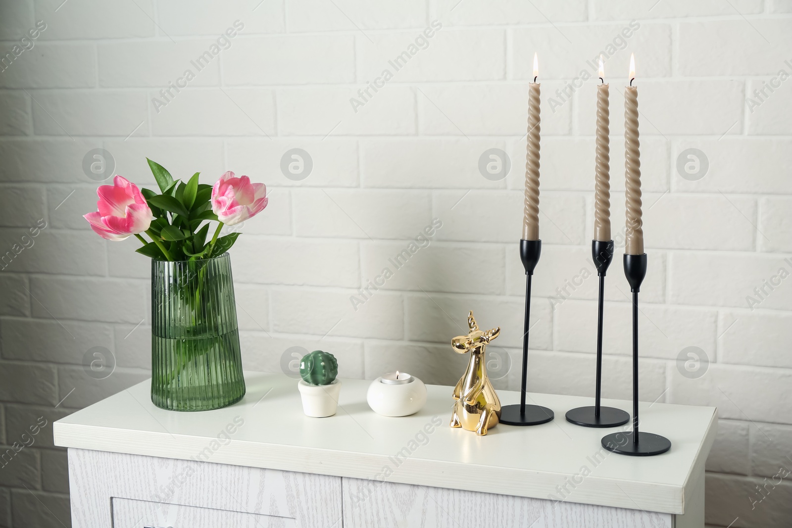 Elegant candlesticks with burning candles, tulips in vase and other decor on dresser near white brick wall Photo of Elegant candlesticks with burning candles, tulips in vase and other decor on dresser near white brick wall