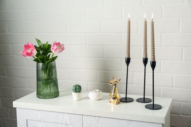 Photo of Elegant candlesticks with burning candles, tulips in vase and other decor on dresser near white brick wall