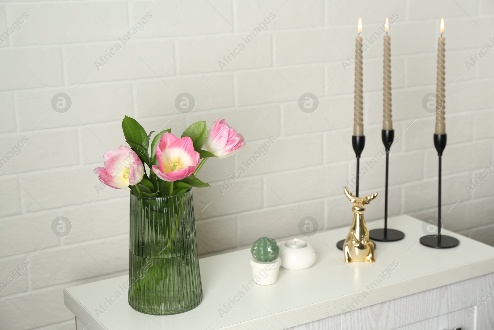 Elegant candlesticks with burning candles, tulips in vase and other decor on dresser near white brick wall Photo of Elegant candlesticks with burning candles, tulips in vase and other decor on dresser near white brick wall