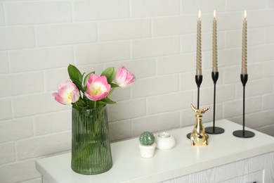 Photo of Elegant candlesticks with burning candles, tulips in vase and other decor on dresser near white brick wall