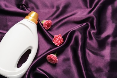 Photo of Bottle of fabric softener and flowers on purple cloth, flat lay. Space for text