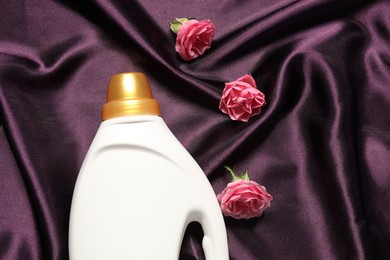 Photo of Bottle of fabric softener and flowers on purple cloth, flat lay