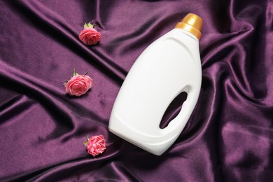Photo of Bottle of fabric softener and flowers on purple cloth, flat lay